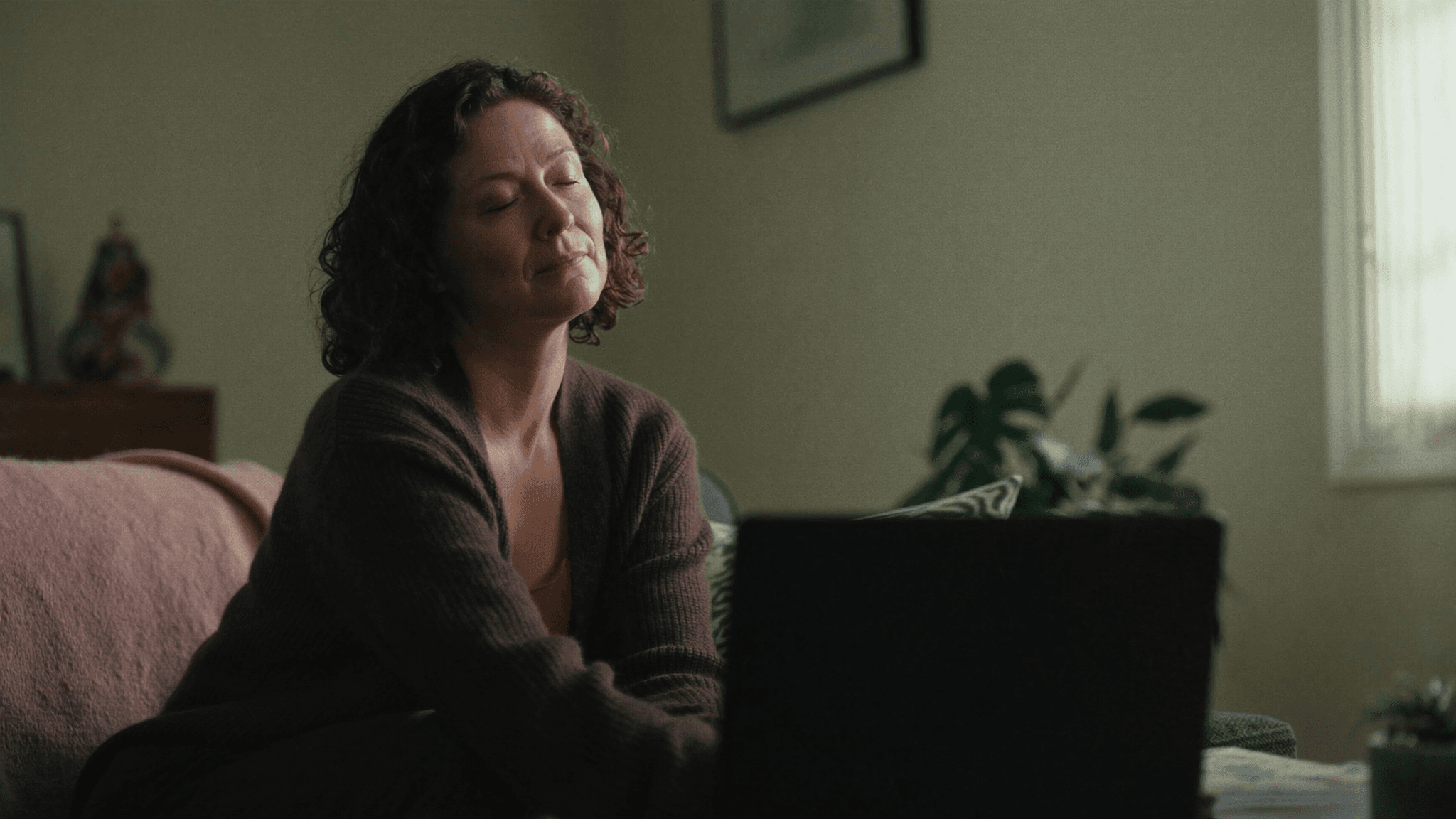 Relaxed woman with eyes closed on a sofa beside a laptop during an online hypnotherapy session at home